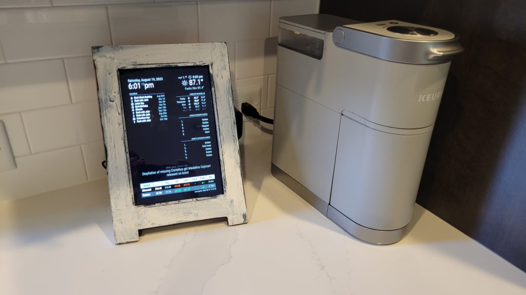 Finished Magic Mirror daily dashboard in a handmade white frame sitting on a countertop beside a coffee maker