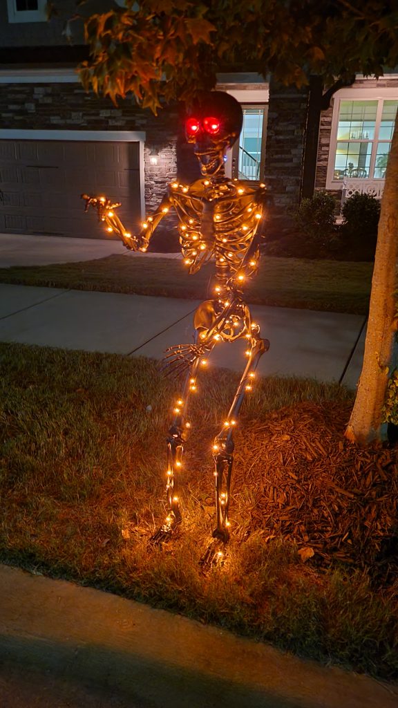 Matte black Halloween skeleton standing in the front yard at night with red eyes and warm light strips glowing