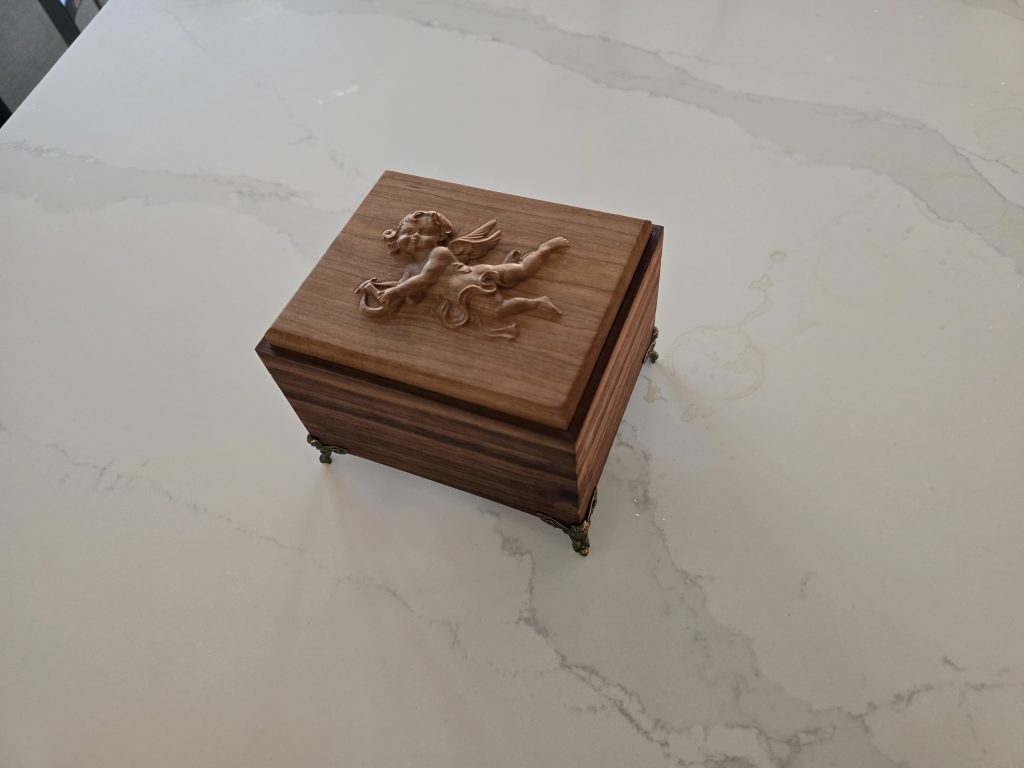 Finished walnut gift box with a cherry cherub lid and metal feet on a countertop.